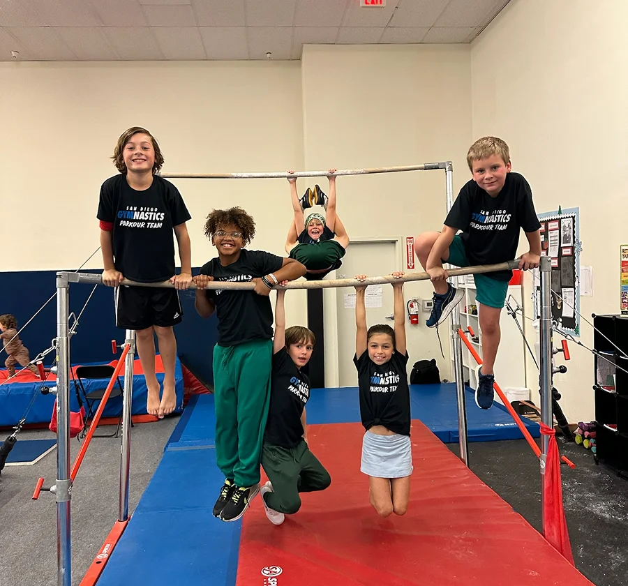 Six children, wearing gymnastics team shirts, smile and pose on parallel bars in a gym. They convey excitement and teamwork in a bright setting.